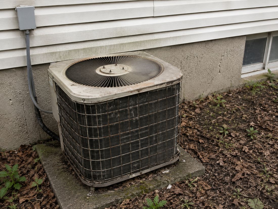 A weathered, rusting outdoor air conditioning condenser on a cracked concrete pad beside a house